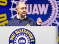 Shawn Fain, a white man with glasses, speaks at a podium. Behind him (and on the podium) is the UAW logo in white on a blue background. Yellow letters around it say (partly obscured) "Our Generation's Defining Moment."