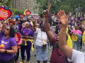 A large crowd of people, mostly Black women, rallies in Manhattan's Foley Square. Many wear purple 1199 T-shirts. Some hold yellow 1199 signs, or red ones shaped like hearts with the message "We are the heart of New York healthcare." The mood is active, with many arms reaching up into the air and mouths opening to speak, chant, or sing.