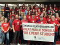 A group of dozens of teachers wearing red shirts gather behind a union banner.