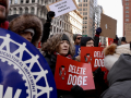 A crowd of people hold red and white signs saying “Delete Doge”