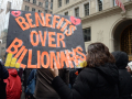 Marchers walk away from the camera with a giant orange on black sign, “Benefits over Billionaires”