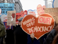 A heart-shaped sign saying “Hands Off Our Democracy” is carried in a crowd.