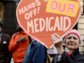 An older person holds up a handmade orange sign saying “Hands off our Medicaid”