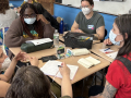 A group of masked people sit around a table discussing something intensely.