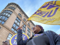 From a heroic low angle, we look up at a Black woman marching in a purple SEIU BJ shirt. The yellow SEIU flag she carries is billowing in the wind, mirroring the curve of the building behind her, against the blue sky. We can see the corners of other picket signs--she is part of a dense crowd.