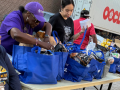 Three women work around a table with blue cloth grocery bags, outdoors. A man in a red T-shirt walks behind them. A truck is also visible.