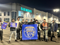 Siemens workers pose in front of the factory in the dark behind a blue banner that says Siemens Workers United.